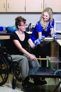 Student helps a woman in a wheelchair to put a casserole dish in the oven.
